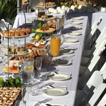 long breakfast table with white tablecloth and elegant dinnerware and food platters in the middle for a birthday party